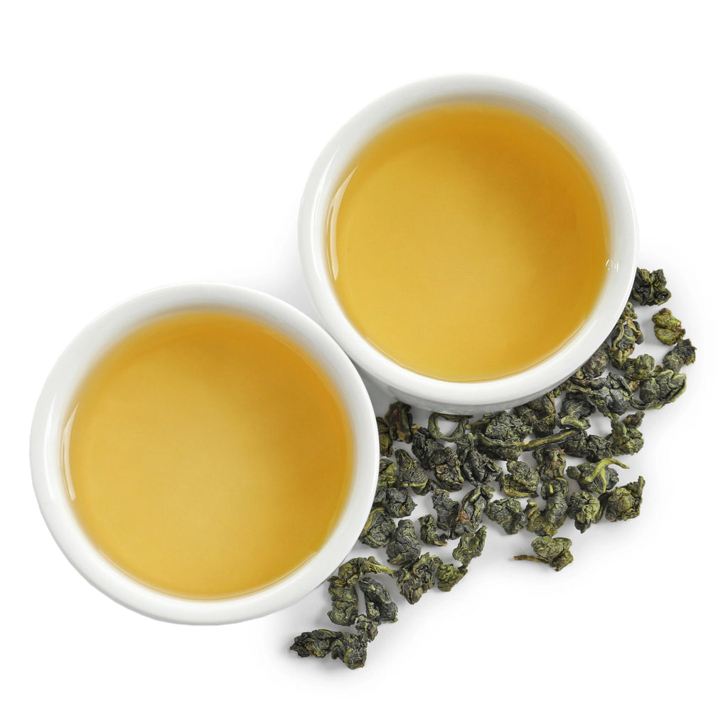 Two white cups filled with yellow tea and a scattering of tie guan yin tea leaves on a white background
