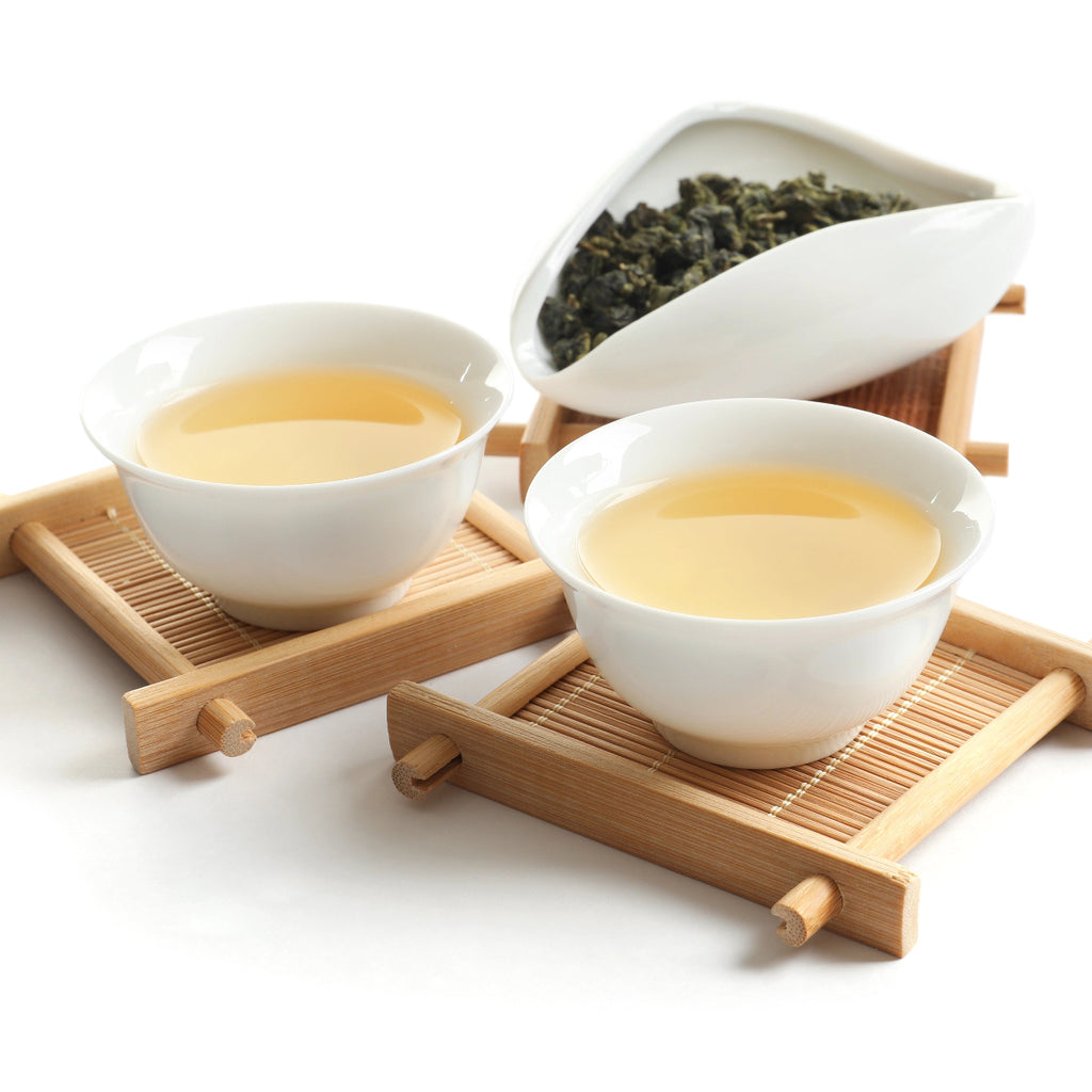 Two white teacups with tea on a wooden coaster, accompanied by a white container of tie guan yin tea leaves.